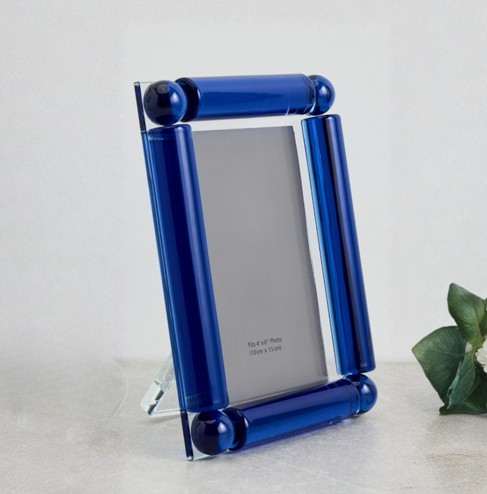 The Tizo Corner Ball Crystal Frame Blue features a modern design with thick, glossy blue glass borders and crystal ball accents. Shown empty on a light surface with green leaves, it comes with a placeholder insert.