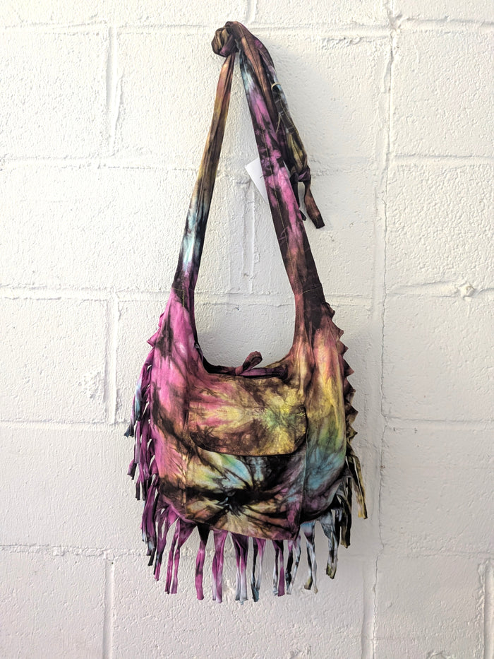 Tie-dye handbag with fringes hanging against a white brick wall.